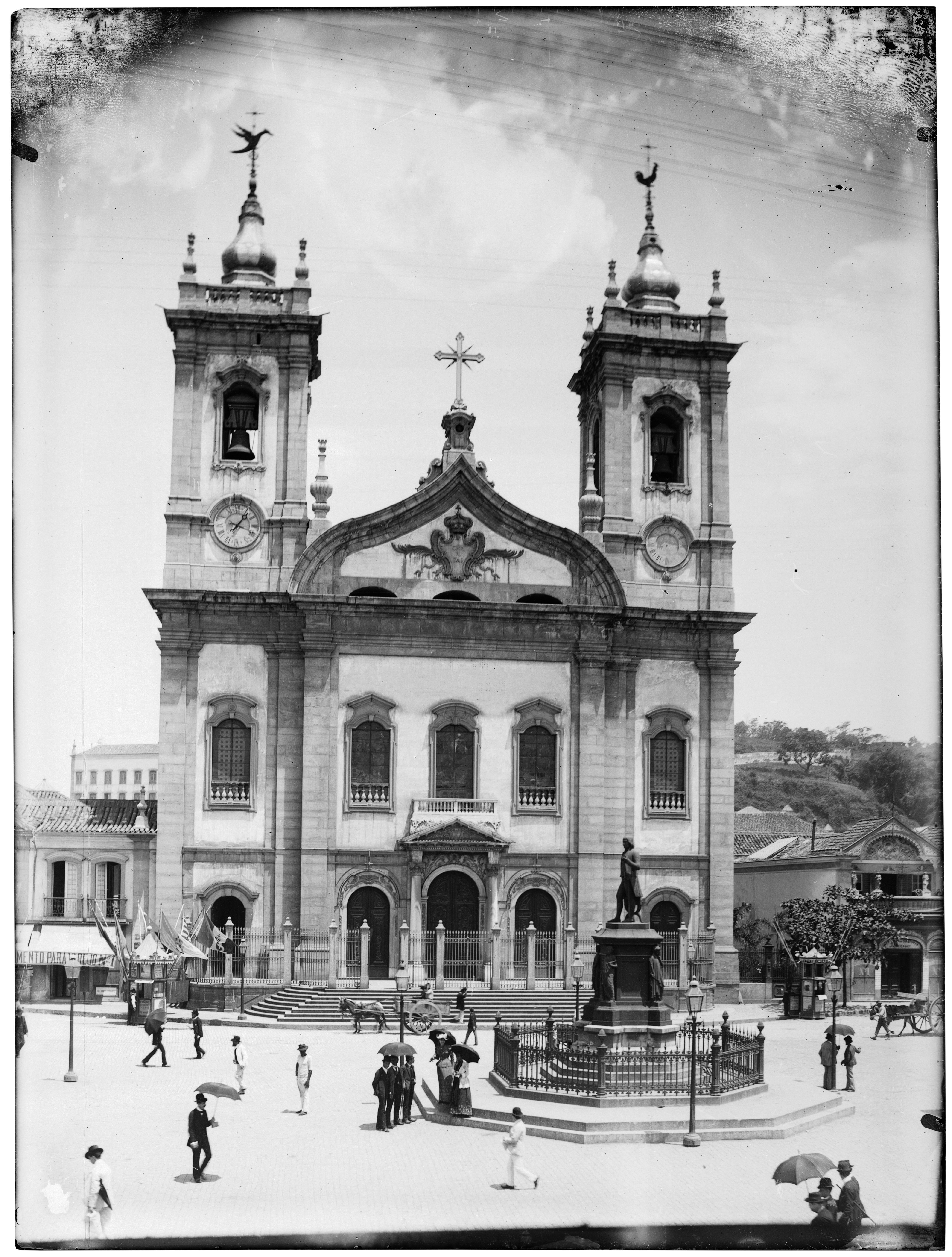 Fotografia de Marc Ferrez, em preto e branco, tirada em 1885, retrata a fachada da Igreja de São Francisco de Paula, no centro do Rio de Janeiro. A igreja possui arquitetura com estilo barroco, duas torres com relógios e uma cruz no meio, além de esculturas religiosas no alto das torres. Em frente ao templo, uma praça movimentada com pessoas caminhando e no centro da praça, um monumento com estátua cercada por grades de ferro. Ao fundo, vê-se árvores, comércios e edificações urbanas.