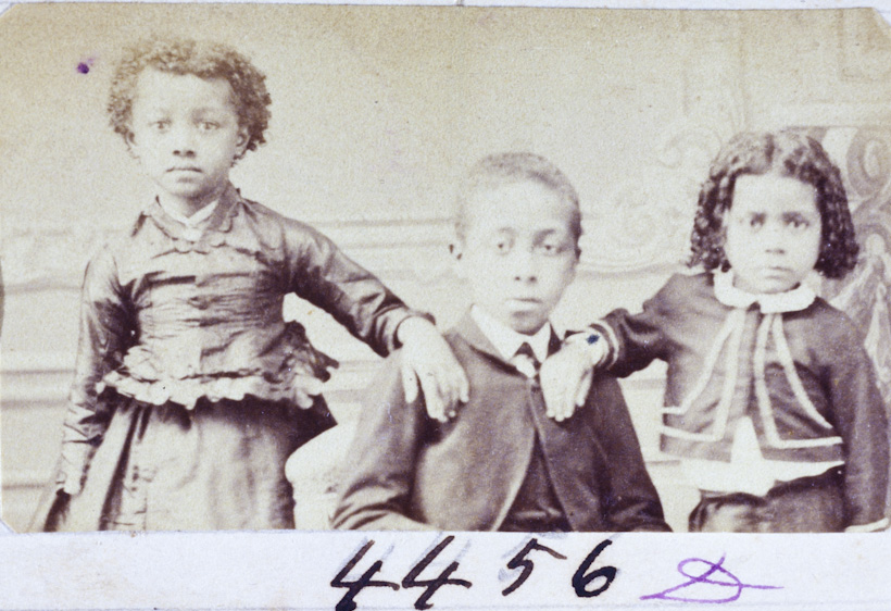 Fotografia do século XIX, em tom sépia, feita por Militão Augusto de Azevedo. A imagem mostra três crianças negras posando diante de um fundo neutro com decoração clássica. À esquerda, uma menina está em pé, com a mão apoiada no ombro do menino ao centro; ela veste um vestido escuro com babados e tem cabelo crespo curto. No centro, sentado, está um menino vestindo paletó, colete e gravata, com expressão séria. À direita, outra menina, também com expressão séria, está sentada com o braço apoiado no ombro do menino; ela tem cabelo encaracolado e veste um conjunto escuro com gola branca e laço. Abaixo da imagem, há a inscrição manuscrita '4456'. A fotografia faz parte do acervo do Museu Paulista da Universidade de São Paulo e documenta com sensibilidade a presença da infância negra na sociedade brasileira oitocentista.