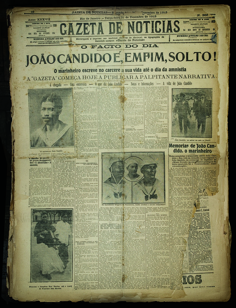 Fotografia de uma página do jornal Gazeta de Notícias, edição de 31 de dezembro de 1912. A diagramação ocupa toda a página com colunas de texto e várias imagens em preto e branco. No topo, em letras garrafais, está o título da matéria principal: 'João Candido é, enfim, solto!', com o subtítulo logo abaixo informando que o marinheiro escreve no cárcere sua vida até o dia da anistia. Abaixo do título há três fotografias em destaque: à esquerda, um retrato de João Cândido com olhar sério; ao centro, uma imagem com três marinheiros fardados; à direita, João Cândido marchando acompanhado por outros homens. Também aparecem imagens menores, como a de duas mulheres. O visual do jornal está amarelado e gasto, com bordas rasgadas e marcas do tempo, evidenciando sua antiguidade. A página transmite a importância histórica do momento, celebrando a libertação de João Cândido após a Revolta da Chibata.