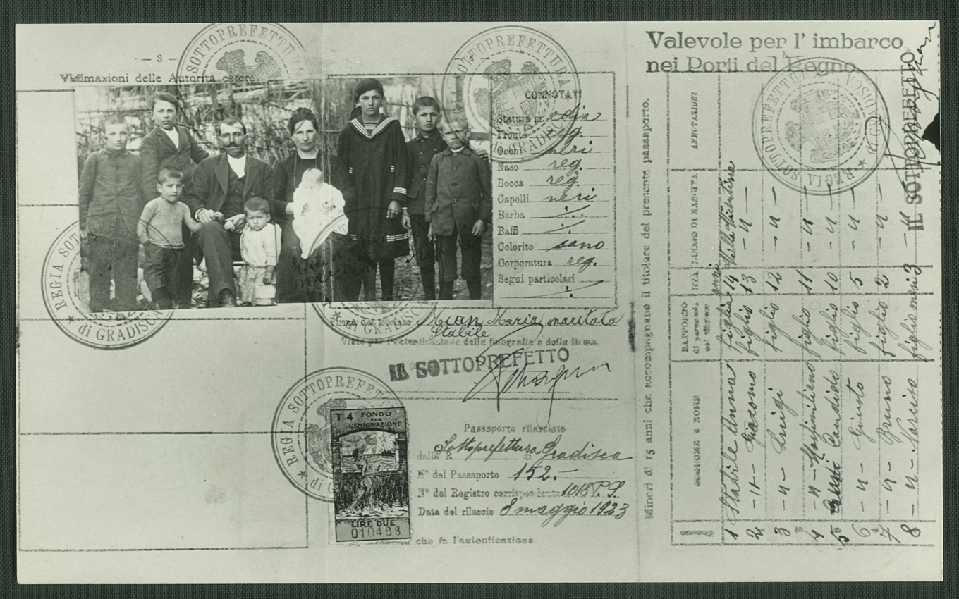 Fotografia de um passaporte italiano de 1923, acervo do Museu da Imigração de São Paulo. O documento, em preto e branco, contém escrita e impressa em italiano, com selos e carimbos oficiais espalhados pela página. No topo direito, há uma fotografia colada de um grupo de imigrantes italianos, entre homens, mulheres e crianças, todos vestidos com roupas de época e postados em posição formal para o registro. O fundo da foto exibe uma paisagem ao ar livre, sugerindo um ambiente rural ou periférico. O passaporte apresenta marcas de envelhecimento, como dobras e leve desgaste nas bordas.