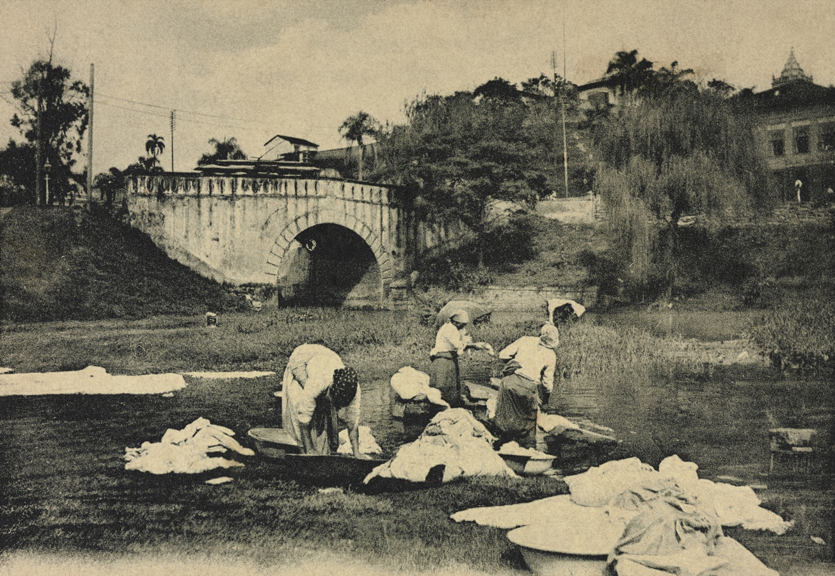 Fotografia em preto e branco intitulada As Lavadeiras às Margens do Tamanduateí, de Guilherme Gaensly, datada entre 1901 e 1905. A imagem mostra um grupo de lavadeiras agachadas e ajoelhadas às margens de um rio, lavando roupas. Peças de tecido estão espalhadas sobre o gramado. Ao fundo, uma ponte de pedra com um arco e, sobre um morro, algumas construções.