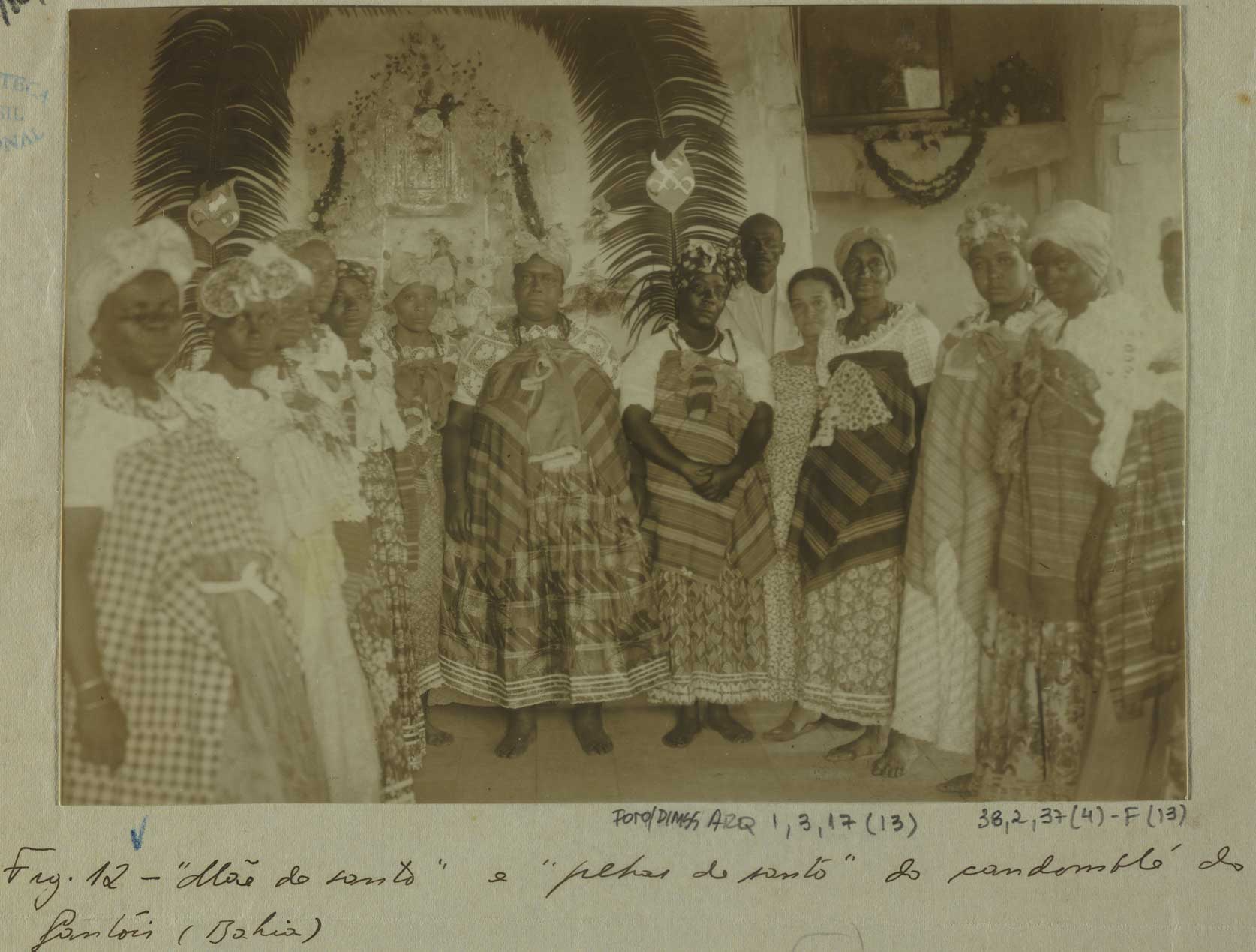 Fotografia/Pintura feita em Salvador (Bahia), em 1940, por Arthur Ramos. Mostra um grupo de mulheres e homens negros reunidos em frente a um altar religioso, decorado com imagens e elementos simbólicos das religiões de matriz africana. Ao centro da imagem está a 'Mãe do santo' — estrutura ornamentada com penas e elementos sagrados, atrás de um conjunto de pessoas com trajes tradicionais do candomblé, como saias rodadas, panos da costa, colares e turbantes. Algumas das mulheres estão de mãos dadas ou lado a lado, formando uma fileira. A imagem representa o contexto das práticas religiosas afro-brasileiras, intensamente perseguidas no início do século XX.